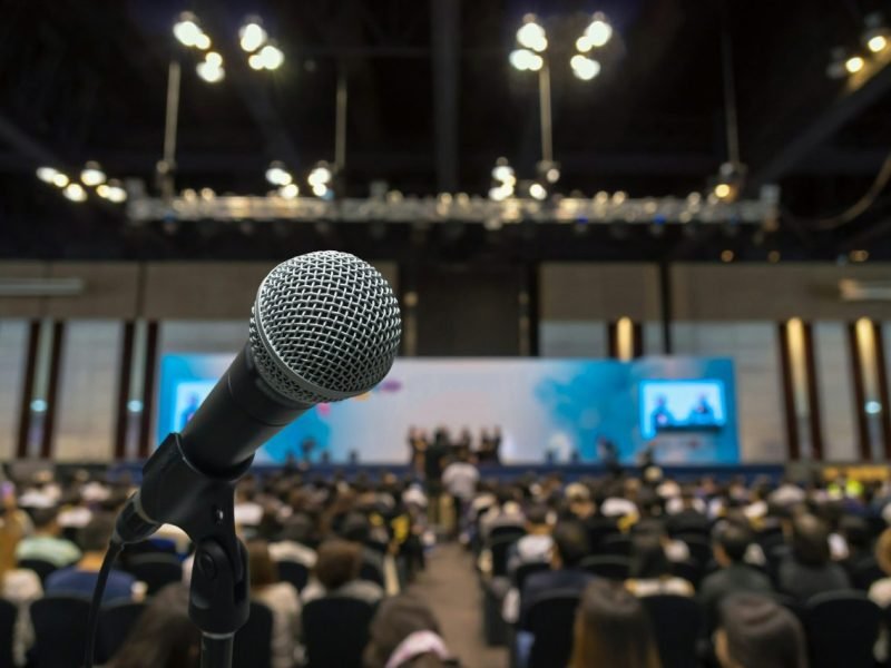 microphone-with-abstract-blurred-photo-of-conference-hall-or-meeting-room-with-attendee-background.jpg